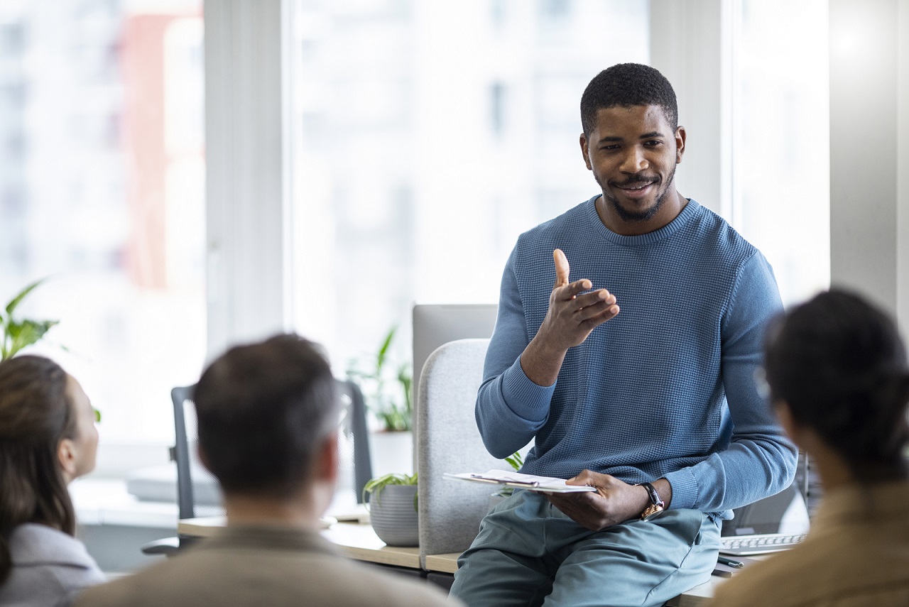 Man leaning on desk talking to people illustrates blog "How To Improve Your Coaching Skills"