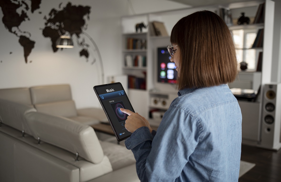 Woman standing in living room holding tablet that reads "Music" illustrates blog "What Is the Internet of Things (IoT)?"