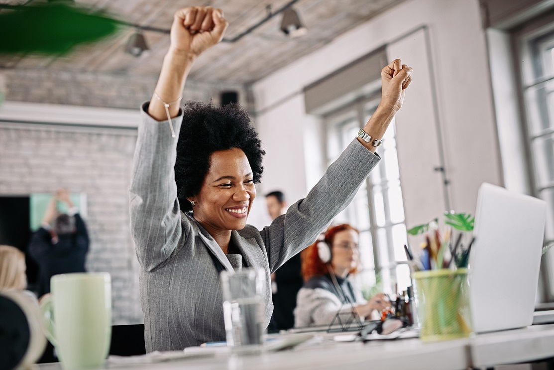 Cheerful African American businesswoman celebrating success while using computer in the office. Motivated employee at desk.